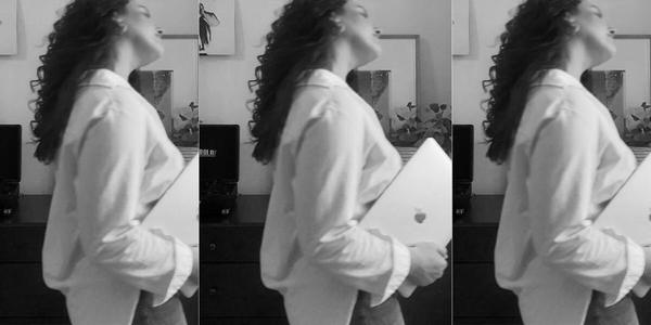 Black and white photo of a side profile of a woman with long, curly hair holding a MacBook in her right hand as she tilts her head to the side